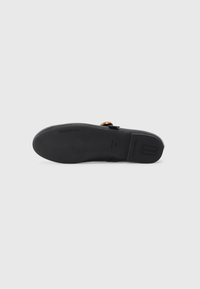 Semelle de chaussure plate noire avec une marque discrète et une boucle dorée visible sur le dessus, sur un fond blanc uni.
