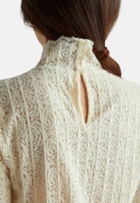 Primo piano della parte posteriore di un top in pizzo crema con motivi floreali verticali e tre bottoni al collo, indossato da una persona con i capelli castani raccolti.