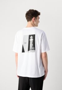 Weißes Baumwoll-T-Shirt mit einem schwarz-weißen Leuchtturm-Motiv auf dem Rücken, kurzen Ärmeln und einem lockeren Schnitt.