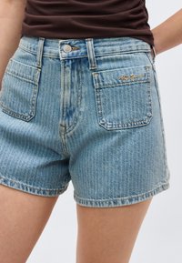 Σορτς από ανοιχτό μπλε τζιν με ψηλή μέση, εμπρός τσέπες με μπαλώματα και καφέ ραφές, φορεμένα με ένα σκούρο καφέ πουκάμισο μέσα από τη φούστα.