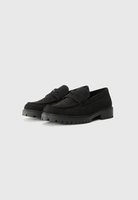 Schwarze Wildleder-slipper mit einer abgerundeten Spitze, dickem Gummisohle und einem dekorativen Riemen über dem Rist. Texturiert und flach für Komfort.