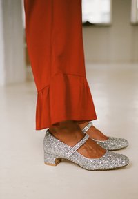 Sidabriniai žvilgantys Mary Jane bateliai su žemu blokiniu kulnu, derinami su raudonais šortais su frakcinėmis lankeliais. Dėmesys blizgiam medžiagos paviršiui ir dizaino detalėms.