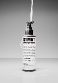 STMTNT-serumflaskan har en svart pumpkork och en vit behållare med fet svart text. Vätska droppar från pipen på en blank yta.