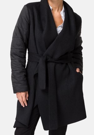 Femme portant un manteau noir ceinturé avec des manches matelassées par-dessus une chemise blanche, main dans la poche, debout devant un fond clair uni.