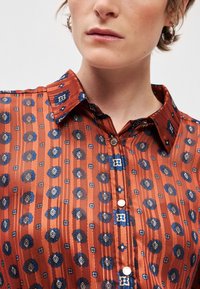 Πουκάμισο με κουμπιά σε καφέ χρώμα με ναυτικές λουλουδάτες και γεωμετρ patterns, που διαθέτει κλασικό γιακά και γυαλιστερά ασημένια κουμπιά κατά μήκος του μπροστινού μέρους.