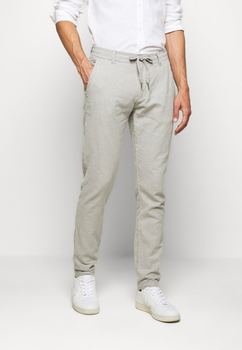 Pantalon en lin gris clair à coupe fuselée, taille avec cordon de serrage et poches latérales. Associé à des baskets blanches sur un fond neutre.