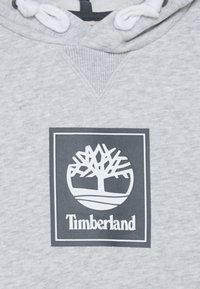 Szara bluza z kapturem z białym logo drzewa w ciemnym prostokącie, z napisem "Timberland" poniżej. Materiał wydaje się miękki i teksturowany.