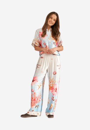 Femme souriante, debout les mains jointes, portant une chemise boutonnée et un pantalon large assortis à motifs floraux avec des dessins de coquillages et de corail.