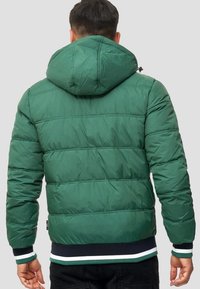 Giacca a vento verde con cappuccio, caratterizzata da sezioni trapuntate, polsini elasticizzati e orlo a bande blu navy con dettagli bianchi.