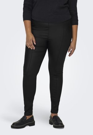 Black, stretchy trousers with a fitted design and seam detailing. Paired with black loafers and a dark top, set against a neutral background.