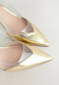Zlaté lodičky na vysokom opätku s elegantným dizajnom, špicatou špičkou a priehľadnými bočnými panelmi, s hladkým, lesklým povrchom.