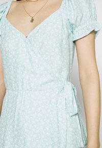 Ljusblå omslagsklänning med blommönster, puffärmar och sidoknytning. Mjukt tyg med en slät yta. V-ringning.