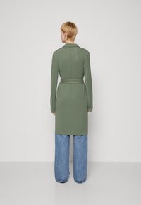 Palton verde lung cu talie cu curea, guler și mâneci lungi, asortat cu pantaloni din denim cu picior larg. Textură netedă cu un design curat.
