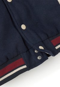 Chaqueta de lana azul marino con grandes botones de presión color crema, puños acanalados con rayas rojas y cremosas, y un tejido de textura suave.