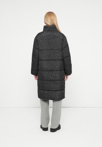Crna puffer jakna s leopard printom, s visokim ovratnikom i horizontalnim quilted dizajnom, uparen sa svijetlosivim hlačama i crnim cipelama.