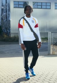 Hvid sportssweatshirt med sorte, røde og gule detaljer, grå crossbody taske, sorte bukser med hvide striber og blå træningssko.