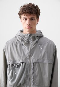 Light gray hooded jacket made of lightweight fabric, featuring a front zipper, adjustable drawstrings, and a mesh pocket with a cinch cord.