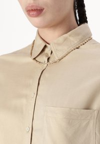 Beige skjorta med knappar och krage, med en bröstficka och slät textur. Förstärkt med ett snurrat guldhalsband.