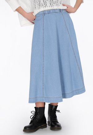 Pull en maille blanche, jupe midi en denim bleu clair avec coutures apparentes, et bottes de combat noires à lacets sur une personne debout.