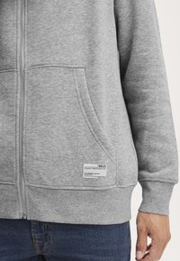 Close-up of a person wearing a light grey zip-up hoodie with front pockets and a small white fabric label, paired with blue jeans.
