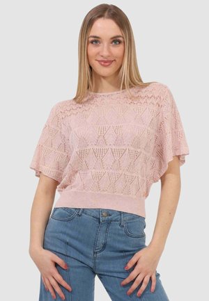 Giovane donna con capelli biondi lisci che indossa una maglietta rosa in pizzo a maniche corte e jeans blu a vita alta, in piedi con le mani vicino ai fianchi.