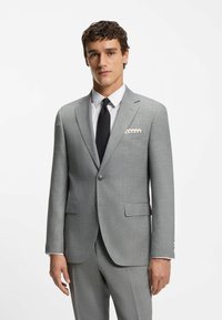 Giacca di suit grigio chiaro con rever a punta, due tasche frontali e tessuto testurizzato. Abbinata a una camicia bianca e una cravatta nera.