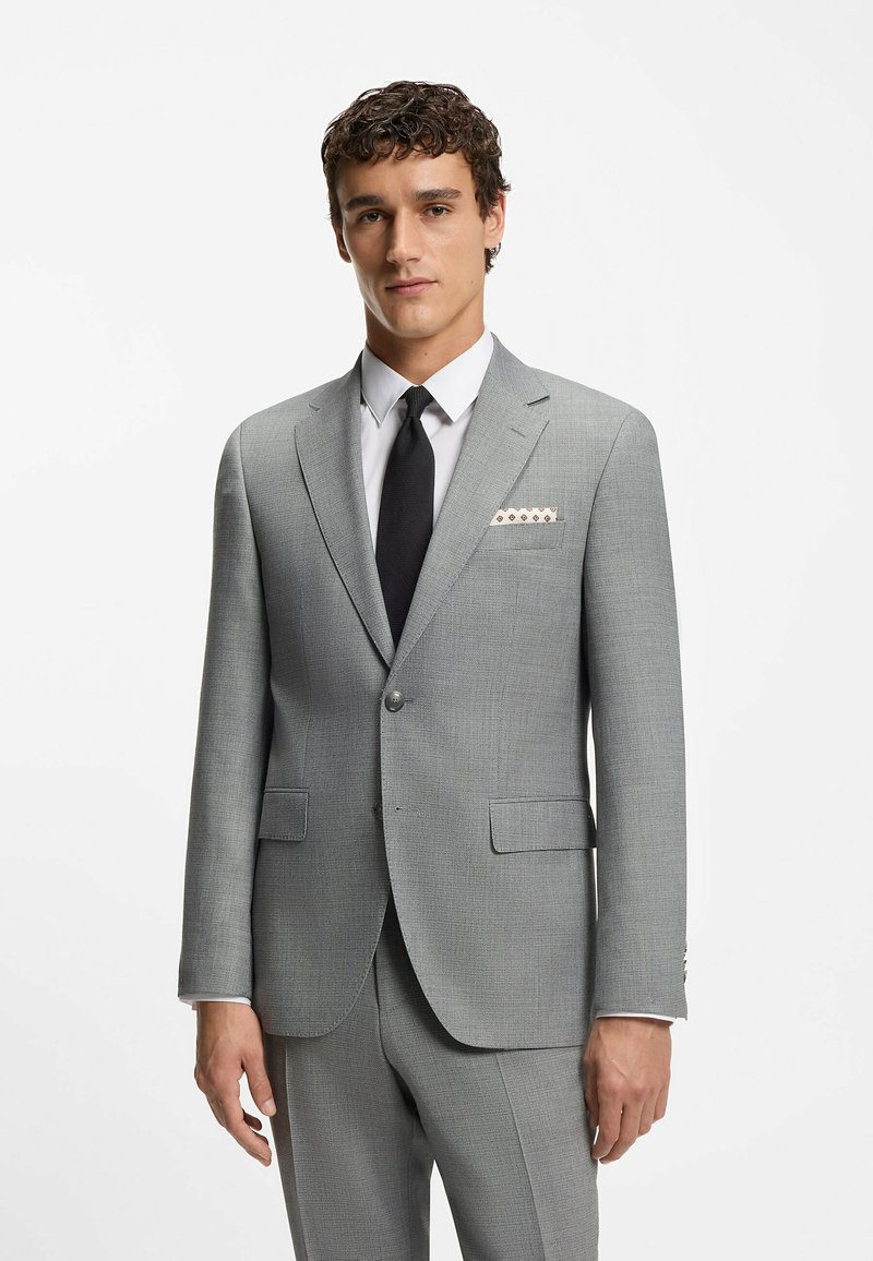 Giacca di suit grigio chiaro con rever a punta, due tasche frontali e tessuto testurizzato. Abbinata a una camicia bianca e una cravatta nera.