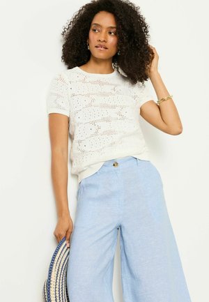 Frau trägt weißes, kurzärmeliges Strickoberteil und hellblaue, weit geschnittene Hose, hält einen gestreiften Hut und berührt ihr lockiges Haar.