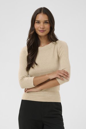 Femeie cu păr lung și castaniu, purtând un top bej cu mâneci lungi și pantaloni negri, stând cu brațele încrucișate pe un fundal simplu.