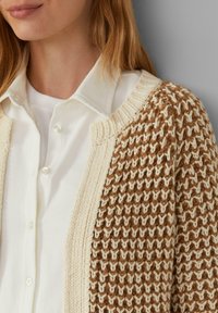 Strickjacke in Creme und Braun mit einem strukturierten Muster. Verfügt über gerippte Kanten und eine lockere Passform, die über einem weißen Hemd getragen wird.