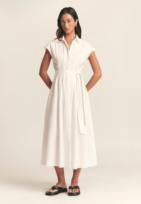 Witte jurk tot op de knie met een nauwsluitend lijfje, kapmouwen en een taillekoord. Lichtgewicht stof met een gladde textuur. Gecombineerd met zwarte sandalen.