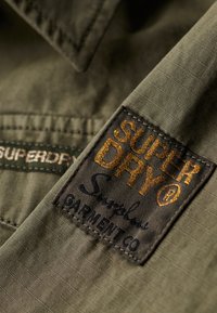 Olijfgroene stoffen shirt met een gestructureerd oppervlak. Bevat een zichtbare Superdry logo-label in goud en zwart met een kenmerkende schrift.