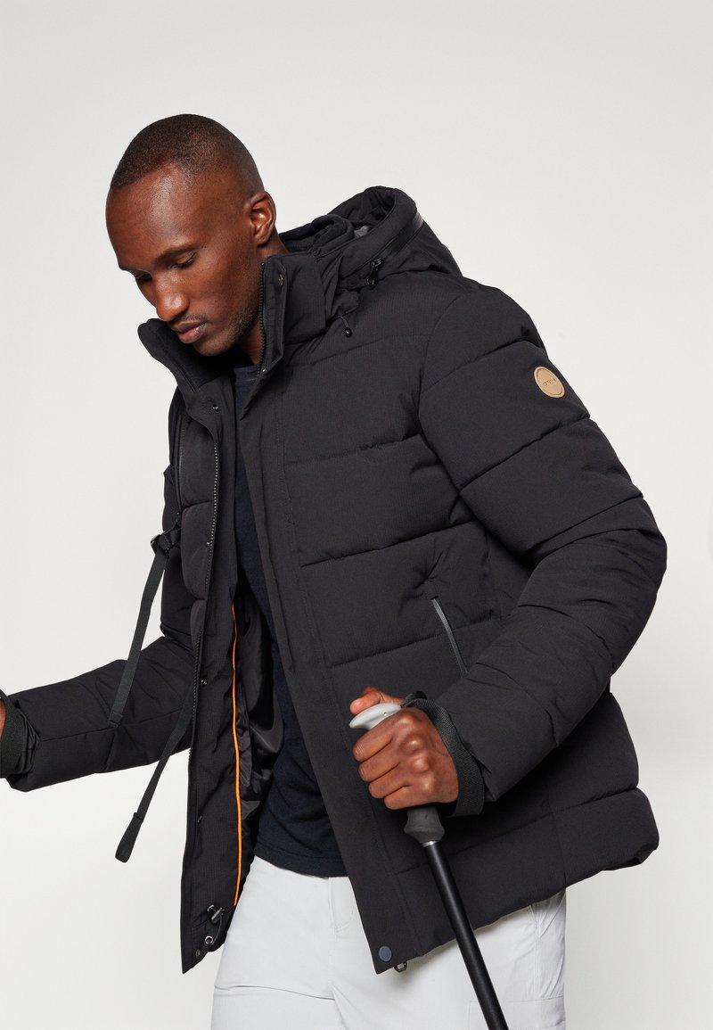 Schwarze Pufferjacke mit strukturiertem äußeren Material, verstellbarem Kapuzen, orangefarbener Innenauskleidung, rundem Logo und Seitentaschen.