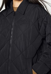 Žena oblečená v černé prošívané bundě se zipem, límcem, vyšitým logem na hrudi a bočními kapsami přes hnědý top.