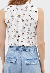 Blusa senza maniche bianca con stampe colorate a tema spiaggia; indossata con pantaloni corti di jeans a vita alta con tasche a pattina.