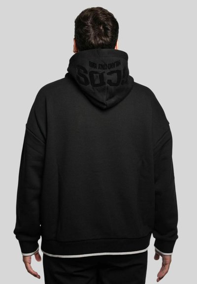 Homme de dos portant un sweat à capuche noir surdimensionné avec du texte sur la capuche, debout devant un fond gris uni.