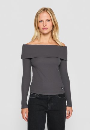 Grauer schulterfreier Oberteil mit langen Ärmeln, geripptem Design und cropped Passform, kombiniert mit dunklen Jeans, die ein sichtbares Logo auf der Tasche haben.