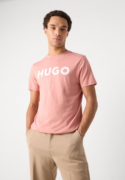Розова памучна тениска с бял логотип "HUGO", с кръгло деколте и къси ръкави, комбинирана с бежови панталони. Гладка текстура.