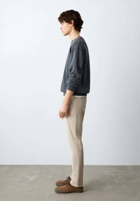 Grauer Pullover, beige Hose, braune Schuhe. Seitenansicht stehend, glatte Textur, schlichtes Design. Neutraler Hintergrund mit hellem grauem Boden.