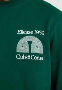 Sudadera verde con un destacado logo bordado en blanco que dice "Ellesse 1959" y "Club di Corsa", junto con un diseño geométrico.