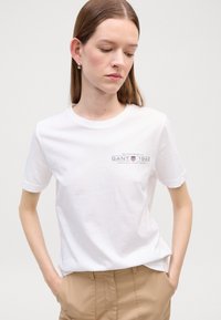 T-shirt bianca di cotone con maniche corte e colletto rotondo, caratterizzata da un piccolo logo stampato in testo scuro sulla parte sinistra del petto.