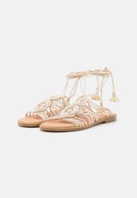 Sandali piatti beige con intricate fibbie intrecciate e legacci alla caviglia che terminano in frange, esposti su uno sfondo bianco.