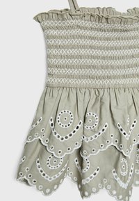 Sytt klänning i ljusbeige tyg med vita broderade mönster. Har en rynkad halsringning och en utfläkt, vågig fåll med utskärningar.