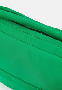 Primo piano di una borsa di tessuto verde parzialmente aperta con cuciture visibili e superficie liscia su uno sfondo bianco.