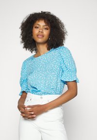 Blusa floral azul clara com mangas curtas volumosas, feita de um tecido macio. Combinada com calças brancas de cintura alta, exibindo uma textura suave.
