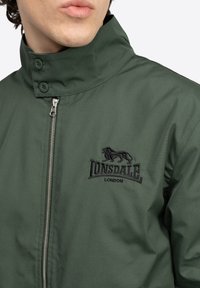 Chaqueta bomber verde con cierre de cremallera, cuello alzado y logotipo negro bordado. Fabricada con un tejido ligero y de textura suave.