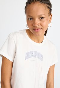 T-shirt en coton couleur crème avec des manches courtes. Présente "NIKE SPORTS" en broderie violette et un petit logo Nike. Texture douce.