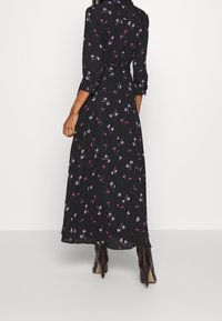 Zwarte maxi-jurk met lange mouwen, bloemenpatroon in roze en paars, taillering met een cinched taille, en luchtige stof, gecombineerd met bruine haklaarzen.