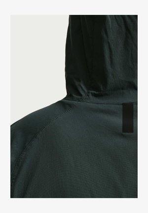 Capuchonjas in donker groene stof. Kenmerken zijn onder andere een gestructureerd oppervlak, een contrasterend zwart label en naden langs de schouders.