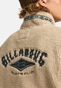 Beige fleecejakke med sort og teal broderet logo på ryggen. Har en mønstret krave og en tekstureret, blød overflade.